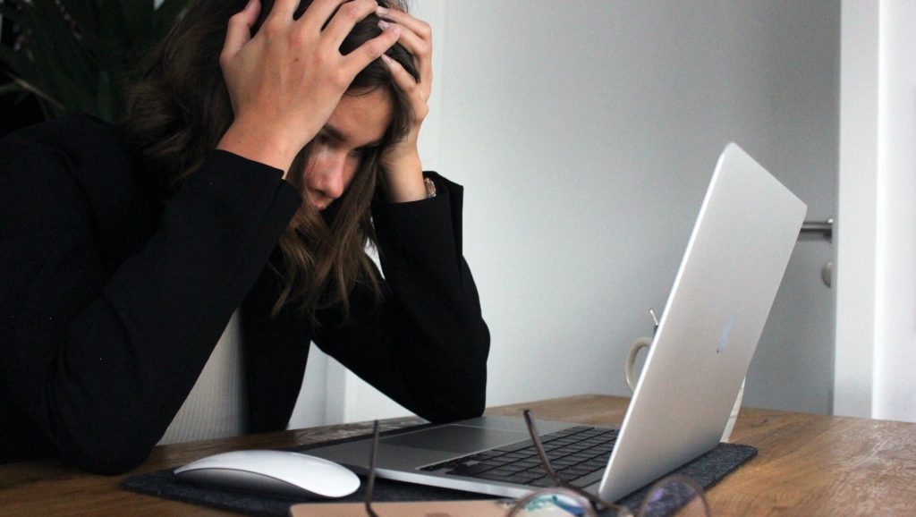 Una mujer sentada en un escritorio, sintiéndose estresada y abrumada mientras se lleva las manos a la cabeza mientras mira la pantalla de su ordenador, reflejando los retos del trabajo o el estudio.