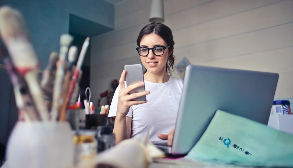 Una mujer sentada en un escritorio, sonriendo a su teléfono mientras su portátil está abierto frente a ella, lo que indica un ambiente de trabajo positivo y productivo